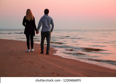 Young couple holding each other hands and walking on sand. Small waves of sea and pink sunset light. Peaceful atmosphere in summer evening. Back view. Empty place for romantic text, quote or sayings.