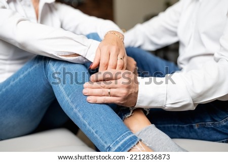 Young couple holding hands with entwined fingers on their engagement day. Polished nails on the female hand. Both are wearing knees. Hands are resting on the knees.