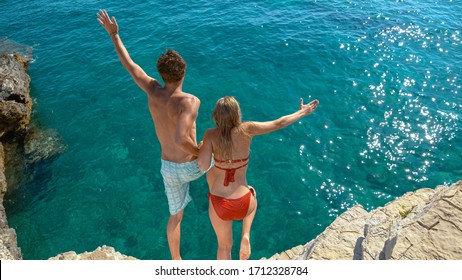 Young Couple Holding Hands Jumps Off A Cliff And Into The Refreshing Cold Ocean. Cheerful Male And Female Tourists Hold Hands While Diving Into Deep Blue Sea. Carefree Man And Woman Jump Into Ocean.