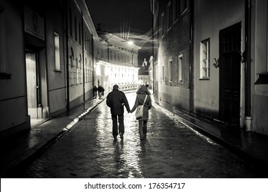 A Young Couple Holding Hands Walking At Night In Old Town Street, Cobbled Street Wet From Rain, Autumn Evening, Black And White. Visible Only Silhouettes Of Models, There Is No Need Model Release.