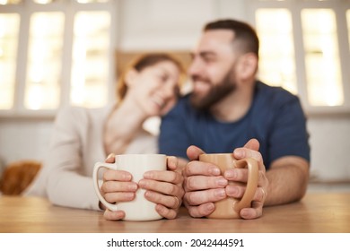 Young Couple Holding Mugs With Both Hands While Leaning On Table In Kitchen