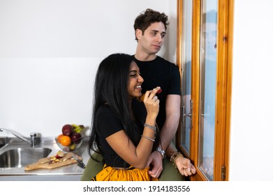 Young Couple At Home Eating Apple In The Kitchen. Indian Woman With Traditional Dress Smiling To Caucasian Man Sitting On Counter Looking Out Window. Lifestyle, Healthy Eating, Multiethnic Concept.