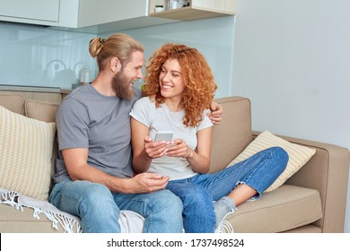 Young Couple At Home Relationship Concept Sitting At Sofa At Living Room Girlfriend Showing Boyfriend Funny Meme On Smartphone Laughing Together Cheerful