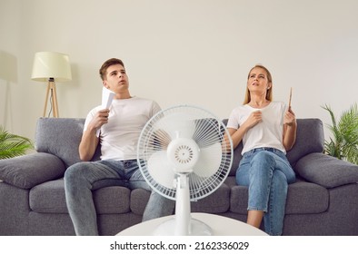 Young couple at home suffering from hot sultry sweltering summertime weather. Sweaty exhausted people waving sheets of paper sitting on sofa with electric fan in overheated house. Summer heat concept - Shutterstock ID 2162336029