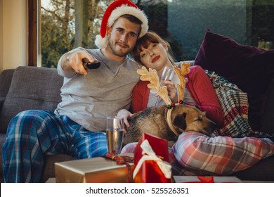 Young couple at home with their pet dog at Christmas time. They are all cuddled up on the sofa at home, watching television. 