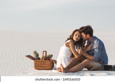 young couple hugging while sitting on blanket with basket of fruits and acoustic guitar on beach - Powered by Shutterstock