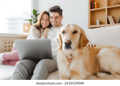 Young couple, husband and wife hugging dog, golden retriever, selective focus using laptop online shopping, sitting on sofa at home. Smiling girlfriend and boyfriend watching video. Technology concept - Powered by Shutterstock