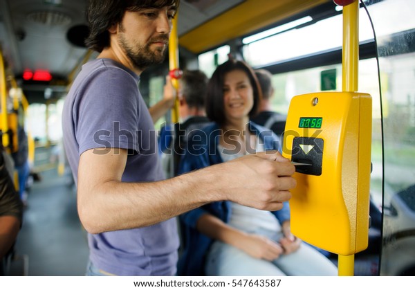Young couple in inside of the city bus. The\
girl sits and transfers to the guy tickets. The young man inserts\
the ticket into the\
validator.