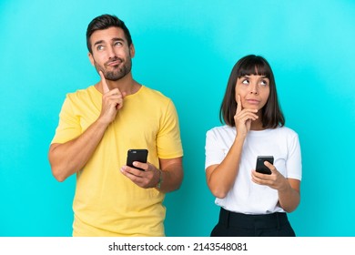 Young Couple Isolated On Blue Background Using Mobile Phone And Thinking