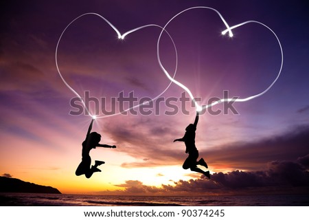 young couple jumping and drawing connected hearts by flashlight in the air on the beach before sunrise