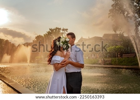 Young couple kisses covered by bouquet of flowers. Couple in love against the backdrop of fountain and splashes. Newlyweds in bright splashes of fountains
