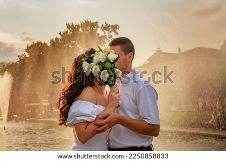 Young couple kisses covered by bouquet of flowers. Couple in love against backdrop of fountain and splashes. Newlyweds in bright splashes of fountains