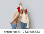 Young couple kissing under mistletoe branch on light background