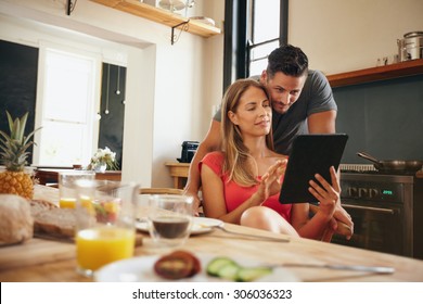 Young couple in kitchen looking at tablet pc. Man standing by his girlfriend sitting using digital tablet in morning. - Powered by Shutterstock