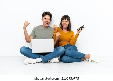 Young couple with a laptop and mobile sitting on the floor celebrating a victory