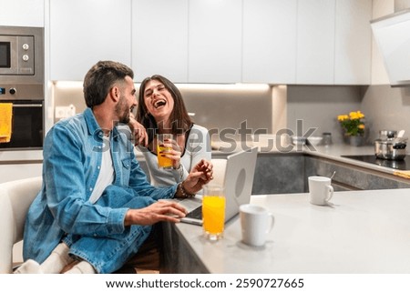 Young couple laughing while using laptop and drinking orange juice in their modern kitchen