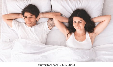 A young couple lies in bed together, seemingly after just waking up. They are both smiling and look relaxed and content. They are wearing casual clothes and are lying on white sheets and pillows. - Powered by Shutterstock