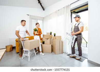 Young Couple In The Living Room Of Their New Home, Mover Performs Professional Furniture Delivery During The Relocation