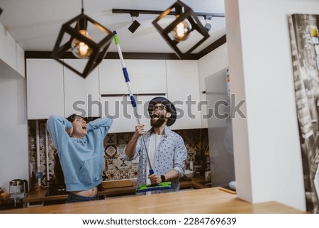 A young couple is looking at the ceiling and screaming while holding a mop because the upstairs neighbor is having a party with loud music or doing repairs in the apartment, workers are drilling with 