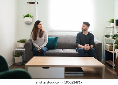 Young Couple Looking At Each Other Angrily While Sitting Apart In The Living Room Sofa After Having An Ugly Fight Because Of Their Relationship Problems