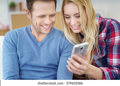 Young Couple Looking At The Screen Of A Mobile Phone Together With Happy Smiles As They Relax At Home On The Sofa