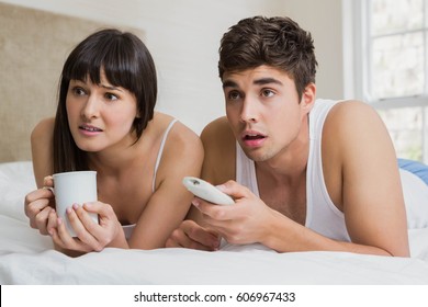 Young couple looking surprised by watching television together on bed in bedroom
