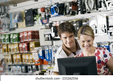 Young Couple Looking At Visual Screen Beside Bike Accessories Display Rack In Bicycle Shop, Smiling