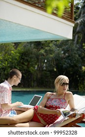 Young Couple Lounging By The Pool In A Garden, Using Technology And Reading A Magazine.