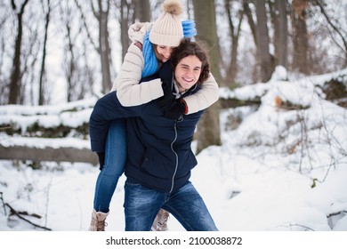 Young Couple In Love Being Goofy And Fun Outdoors In The Forest During Winter. Man Carries The Woman On His Back, Piggyback Style.