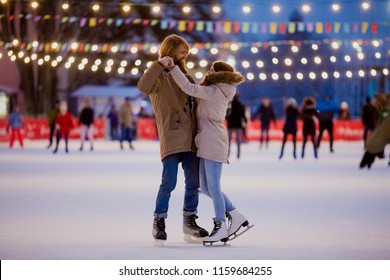 Young couple in love Caucasian man with blond hair with long hair and beard and beautiful woman have fun, active date ice skating on the ice arena in the evening city square in winter on Christmas Eve