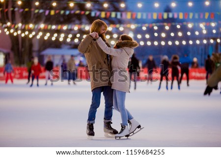 Young couple in love Caucasian man with blond hair with long hair and beard and beautiful woman have fun, active date ice skating on the ice arena in the evening city square in winter on Christmas Eve