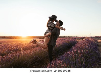 A young couple in love celebrates their anniversary with a romantic date in a lavender field at sunset. The guy picks up his girlfriend in his arms, looking at each other and smiling. - Powered by Shutterstock