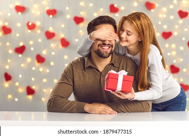 Young couple in love celebrating Saint Valentine's Day or relationship anniversary. Happy woman covering boyfriend's eyes giving him surprise gift. Smiling man getting present from loving girlfriend - Shutterstock ID 1879477315