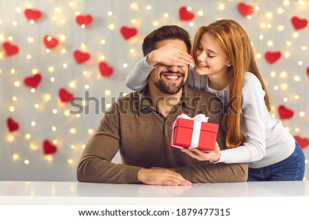 Young couple in love celebrating Saint Valentine's Day or relationship anniversary. Happy woman covering boyfriend's eyes giving him surprise gift. Smiling man getting present from loving girlfriend