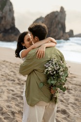 Young Couple In Love Embracing Having Romantic Date On Beach, Man Holding Flowers Behind His Back, Giving Gift To His Girlfriend, Vertical Shot