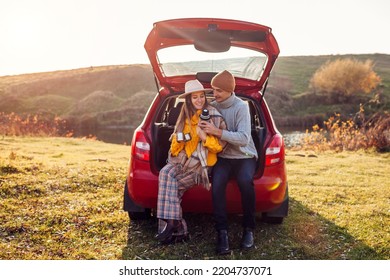 Young couple in love relaxing in car trunk by autumn lake drinking tea from vacuum flask. Man and woman travel by auto. Young man and woman enjoy fall landscape
