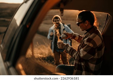 Young couple in love sitting in car open trunk drinking coffee. A male hiker approaches the car, a woman is pouring a fresh coffee from the thermos. - Powered by Shutterstock