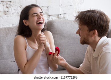 Young couple in love sitting at couch at home. Smiling man holding red box with marriage engagement ring got up on his knee and make proposal to happy surprised emotional girlfriend. Marry me concept
