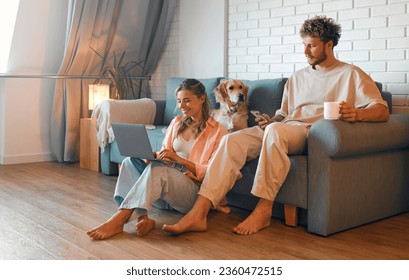 A young couple in love sitting in the living room in a cozy home, using a laptop and phone, sitting on the sofa with their beloved dog and drinking morning coffee. - Powered by Shutterstock