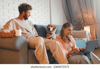 A young couple in love sitting in the living room in a cozy home, using a laptop and phone, sitting on the sofa with their beloved dog and drinking morning coffee. - Powered by Shutterstock