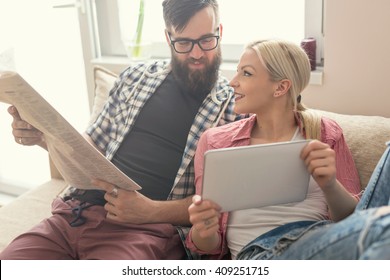 Young Couple In Love Sitting On A Couch In Their Apartment Next To The Window, Enjoying Their Free Time, Reading Newspaper And Surfing The Web On A Tablet Computer. Lens Flare Effect On Window