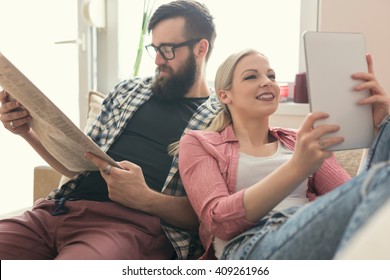 Young Couple In Love Sitting On A Couch In Their Apartment Next To The Window, Enjoying Their Free Time, Reading Newspaper And Surfing The Web On A Tablet Computer. Lens Flare Effect On Window