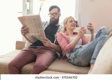 Young Couple In Love Sitting On A Couch In Their Apartment Next To The Window, Enjoying Their Free Time, Reading Newspaper And Surfing The Web On A Tablet Computer. Lens Flare Effect On Window