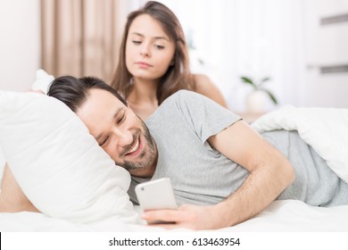 A Young Couple Lying In Bed. One Of Them Use The Phone And Flirting. The Other Person Is Jealous And Spies Of The Arm. The Concept Of Jealousy And Distrust.