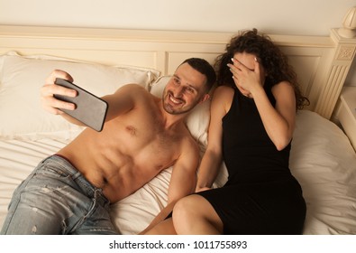 Young Couple Lying In The Bed. Couple Taking Selfie. Woman Is Embarrassed. Happy Couple Photographed