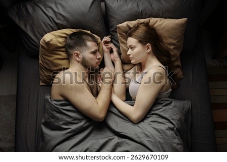 Young couple lying in bed while looking into each other's eyes, with intimate expressions and cozy atmosphere and soft pillows creating relaxing scene