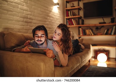 A Young Couple Lying On The Couch As The Boyfriend Is Browsing A Tablet For A Movie To Watch.