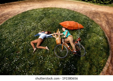 Young Couple Lying On The Grass And Riding The Bicycle With Red Dirigible And Basket With Baguettes In The Park , Top View, General Plan