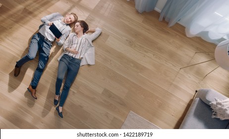 Young Couple Are Lying On A Wooden Flooring In An Apartment. They Are Using A Tablet Computer. Cozy Living Room With Modern Interior, Grey Sofa And Wooden Parquet. Top View Camera Shot.