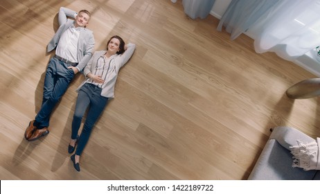 Young Couple are Lying on a Wooden Flooring in an Apartment. They are Happy, Smile and Laugh. Cozy Living Room with Modern Interior, Grey Sofa and Wooden Parquet. Top View Camera Shot.
